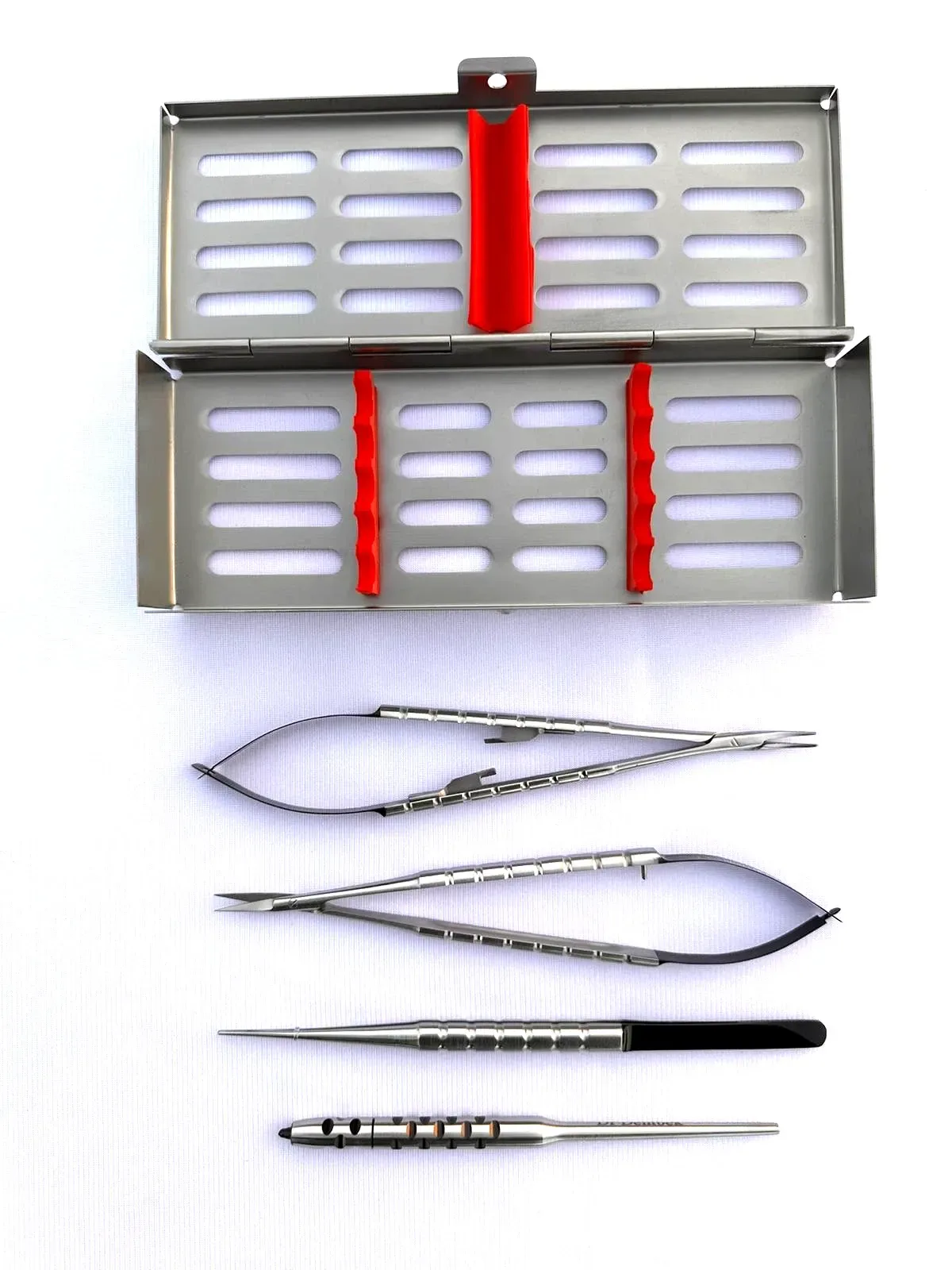 Open, empty metal cassette above four separated instruments: a spring needle holder with jaws to the right, spring scissors with blades to the left, fine tweezers pointing left and a round scalpel handle at the bottom pointing right, with no blade fitted